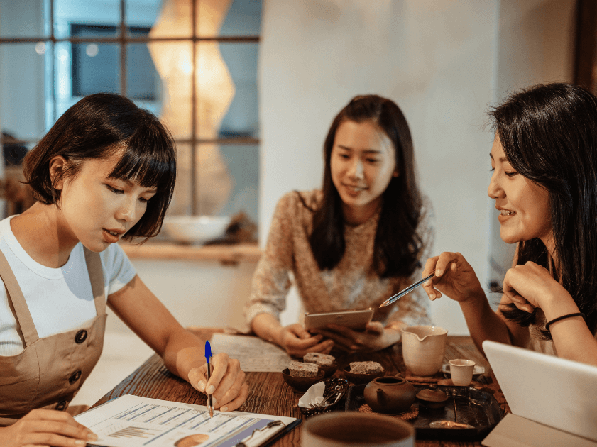 A group of three women sitting round a table, collaborate on a document.