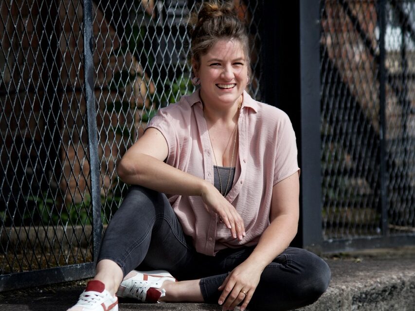 Tilley Bancroft, Founder of Making Trails. A woman sitting in front of a chain fence, wearing a pink top and black jeans.